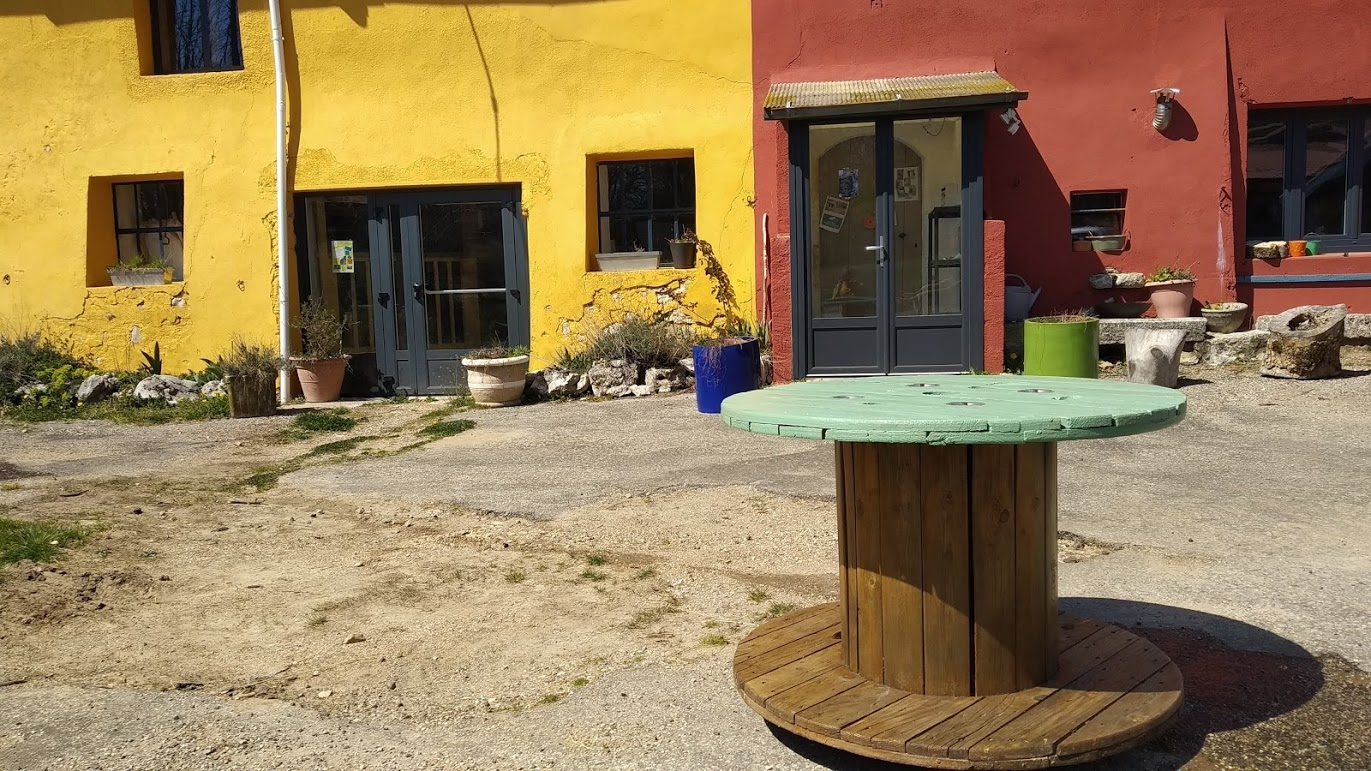 La cour et ses tables en touret devant les façades jaune et rouge, en plein soleil